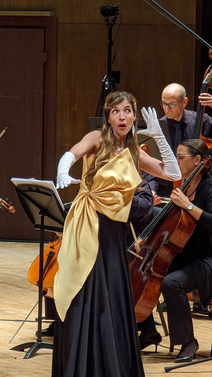 Monica in a black-and-gold gown and white gloves, gesturing beside a music stand with string players behind her.