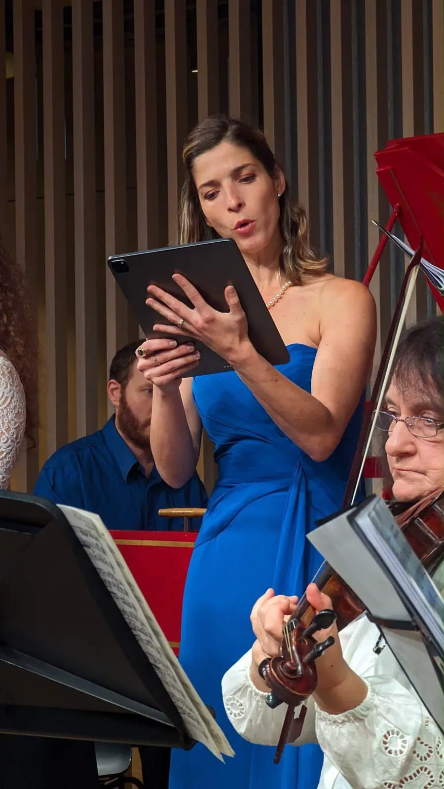 Monica in a blue dress holding a tablet and singing, with a music stand and string players close by.