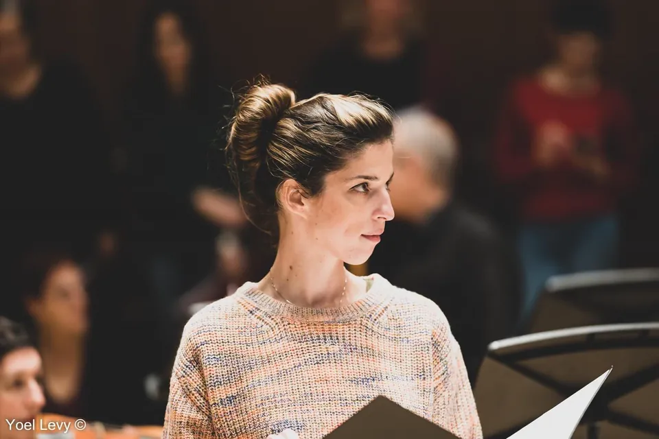 Close-up of Monica in a light multicoloured knitted sweater, holding a dark music folder and looking to the right; seated people are softly blurred behind. The photographer’s watermark is kept at the lower left.